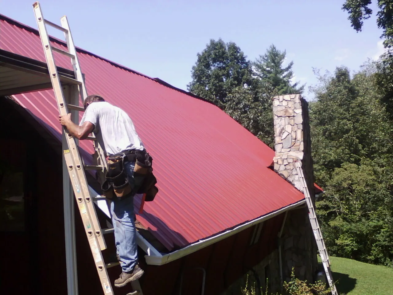 Roofer on a ladder beside a red metal roof and stone chimney — Soffit & Fascia Repair by Roofing Brothers in Mc Louth
